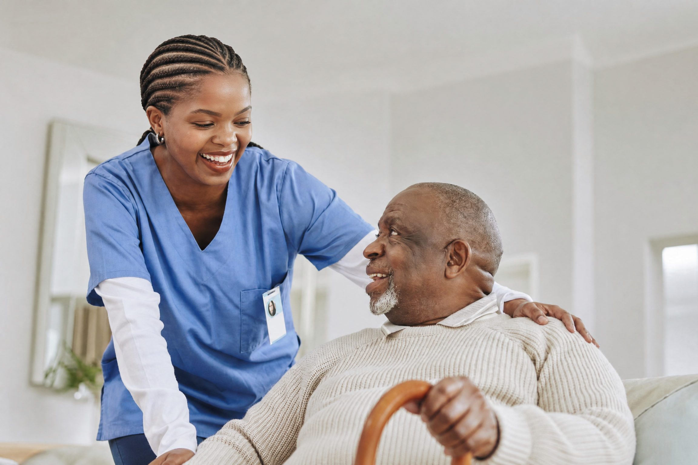 A Black caregiver in blue scrubs smiling with an older man during an in-home care visit.